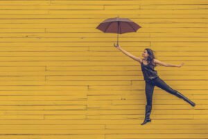 Frau mit Schirm springt in die Luft vor gelber Wand